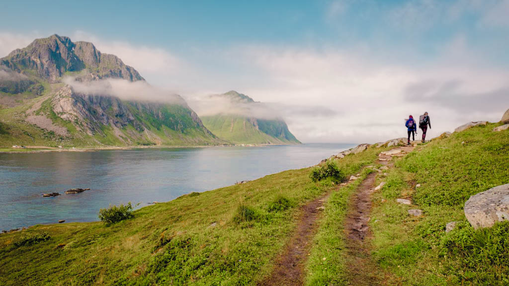 Two hikers on a coastal path in Lofoten, the landscape around them dotted with potential places to stay in this breathtaking natural setting.