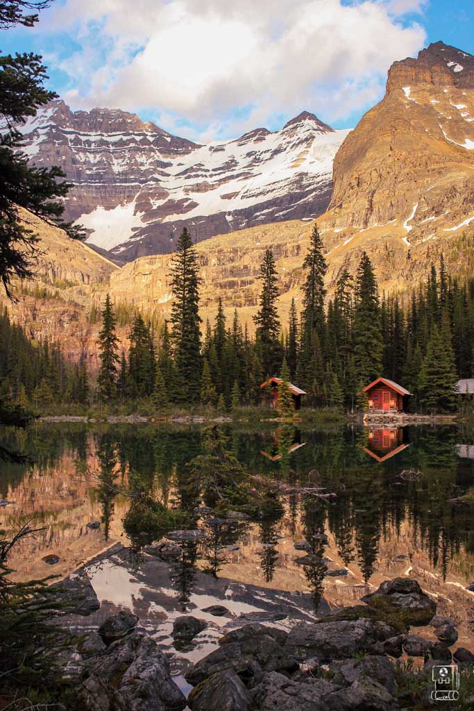 Lake O'Hara Lodge cabins reflecting in the crystal-clear alpine lake at sunset, with snow-capped peaks and evergreen forest creating a perfect endpoint for hiking itineraries